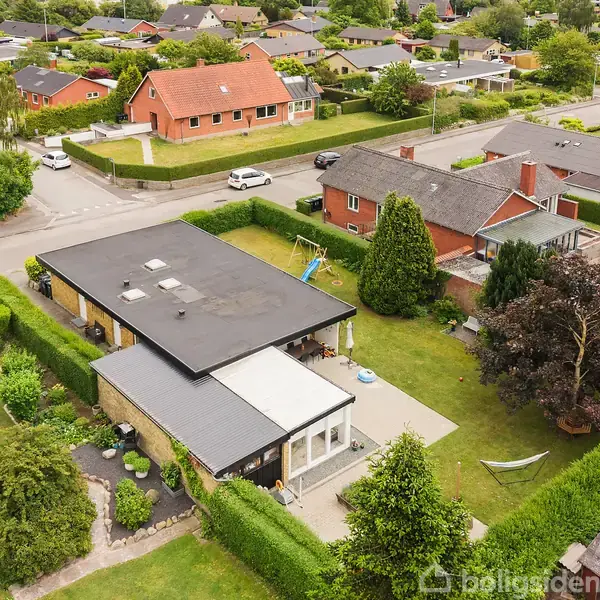 Et moderne hus med fladt tag står i en velplejet have med tæt hæk. Haven har et lille legeområde med gynge og grønne omgivelser. Omkringliggende nabolag med flere huse.