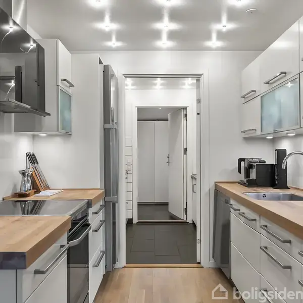 Et moderne køkken med lyse trækøkkenskabe, stænkløse bagplader og en glaskeramisk komfurplade. Køkkenet er lyst op af spotlights. En kaffemaskine står ved siden af vasken.