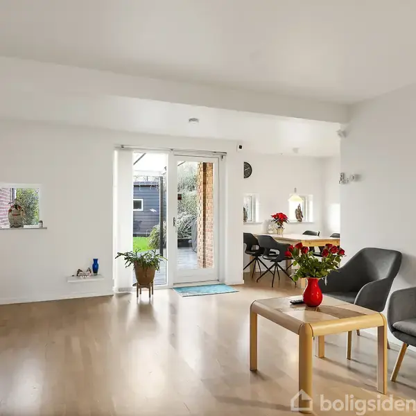 Stue med træbord, grå stole og tv-møbel. Bordpynt og blomster dekorerer rummet. Dør åbner til terrasse med planter. Rummelig og lys indretning med trægulv.