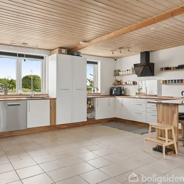 Lyst køkken med hvide skabe, trædetaljer og moderne apparater; kaffemaskine på bord med to høje stole. Hylder med kopper. Store vinduer giver udsigt til grønt landskab. Tekst: “Home Sweet Home”.