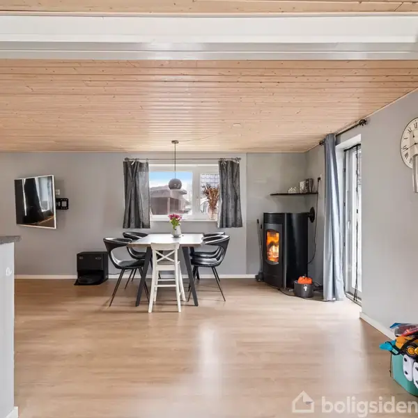 Spisebord med sorte stole står i en lys spisestue. Pejsen brænder ved siden af en stor vinduesdør. Væggene har grå nuancer med et vægur over.