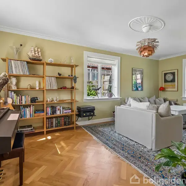 Et stueområde med en grå sofa står på et blomstret tæppe. En reol med bøger og pynt genstande ses ved siden af et klaver. Store vinduer giver lys.