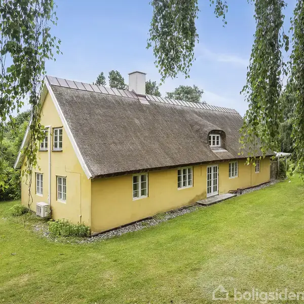 Et stråtækt hus står stille omgivet af en frodig have. Bygningen er malet gul og har flere vinduer på begge etager. Træer og græsplæne danner rammen om huset.