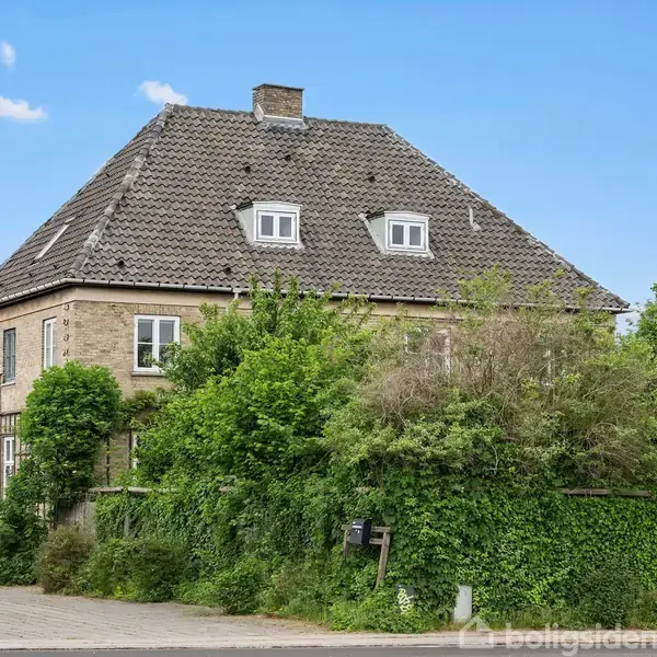 Et stort, firkantet hus med gråt tag og flere små vinduer er omgivet af frodige træer og buske. Huset står i en grøn, velholdt have på en rolig vej.