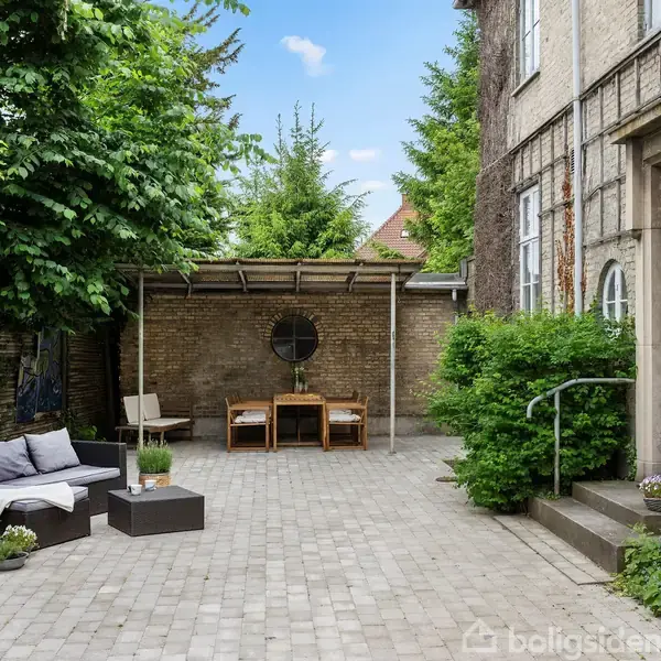Stole og sofa står på en brostensbelagt gårdhave foran en bygning med blå dør. Et bord og stole er placeret under en overdækning ved en mur med runde vinduer og grønne planter.