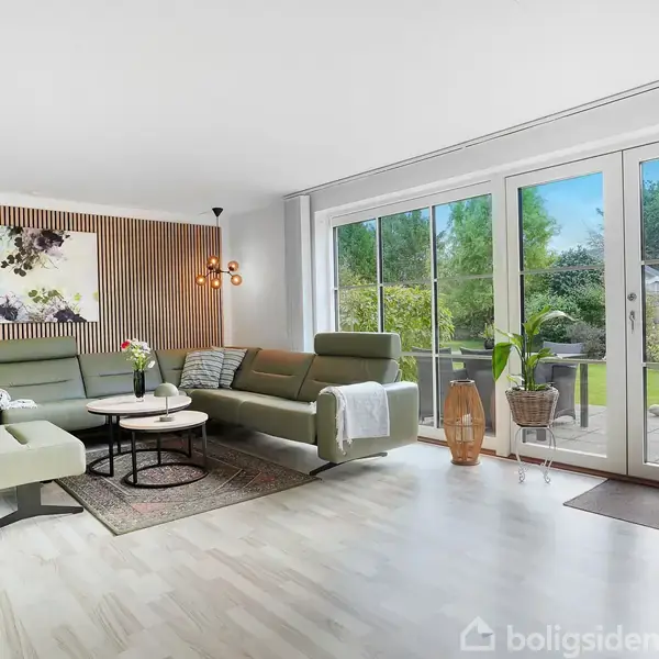 En moderne stue med grønne sofaer står foran store glasdøre, der viser en have udenfor. En plante, et kunstværk, og en hængelampe pynter rummet.