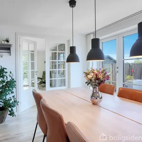 Et spisebord med brune stole står i centrum. Bordet er pyntet med en blomsterbuket og en lyserød lysestage. Tre sorte lamper hænger over bordet. Store vinduer og en åben dør ses i baggrunden.