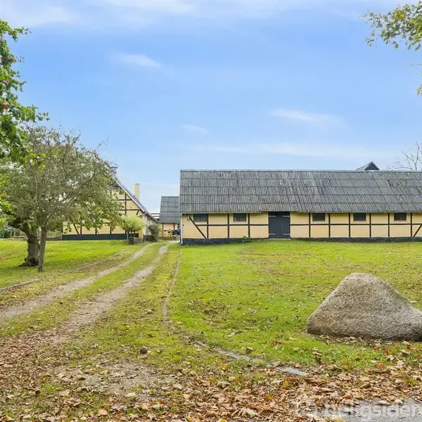 Gårdbygning med sort bindingsværk står stille omgivet af græs og træer. En grusvej fører op til bygningen, der præsenterer en landlig atmosfære under en klar blå himmel.