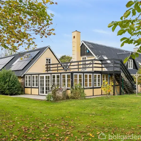 Hus med sort tag og lysegule vægge, har en altan med udvendig trappe, omgivet af en grøn have med træer.