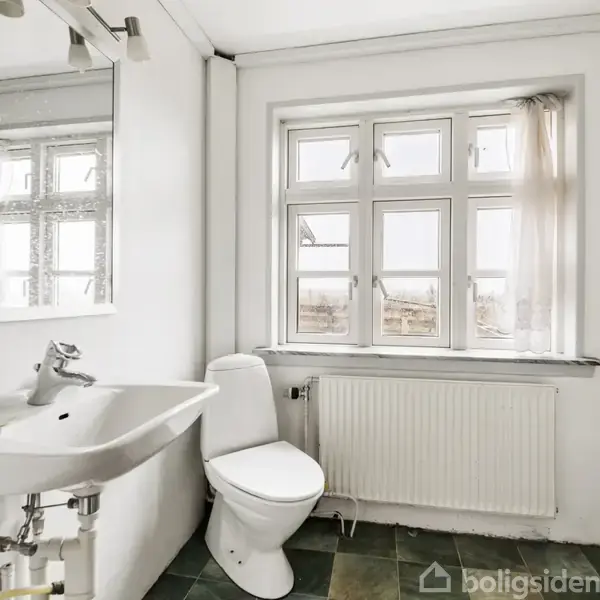 En hvid vask med spejl over hænger på væggen. Ved siden af er et toilet. Rummet har en stor vindueskarm med gardiner og fliser på gulvet.