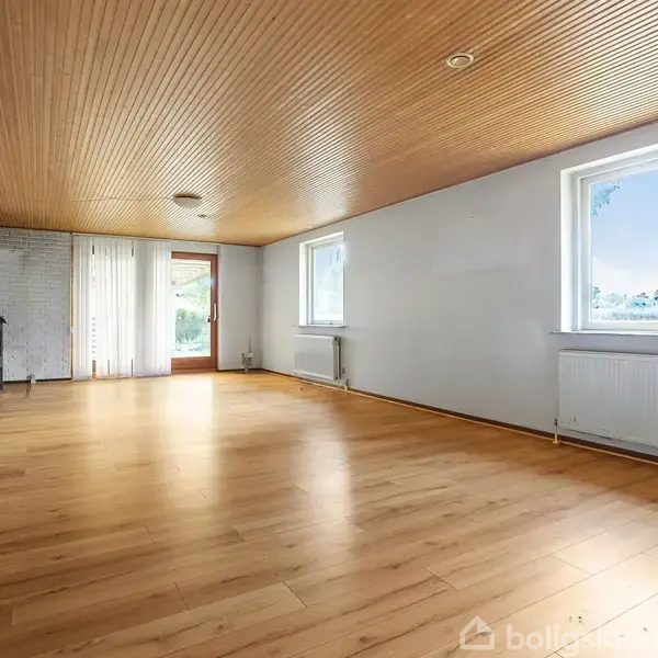 Et rummeligt, tomt rum med trægulv og træloft. En brændeovn står i hjørnet ved en hvid murstensvæg. Store vinduer og en glasdør giver udsigt til naturen udenfor.