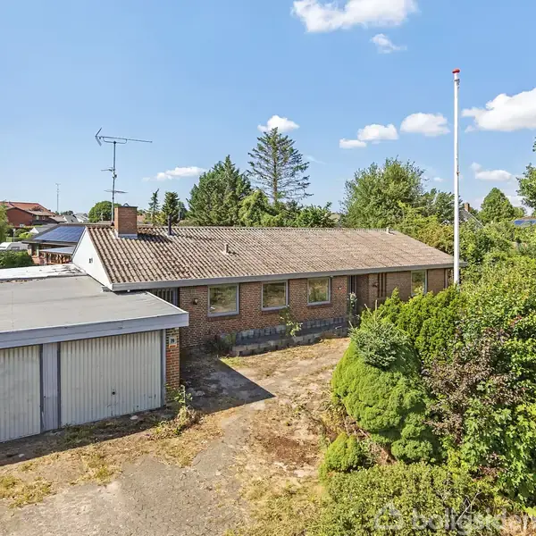 En étplans murstenshus med fladt tag står stille i en villahave. Foran huset er en garage og en flagstang. Haven er frodig med buske og træer omkring.