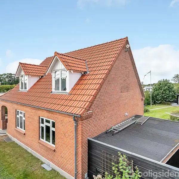 Et rødt murstenshus med rødt tegltag står i en forstad. Huset har to stueetager og kviste på taget. Haven er omkranset af hække og ligger i et grønt område.