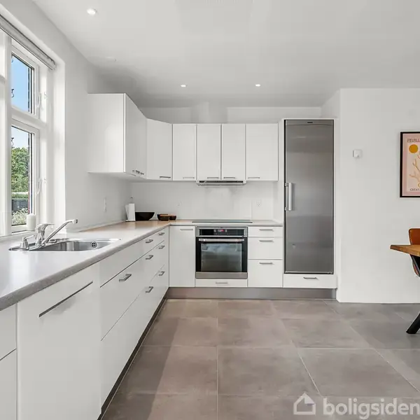 Et lyst køkken med hvide skabe og grå bordplader. Ovn og køleskab er integreret. I baggrunden står et spisebord med to brune stole. Vinduerne viser en grøn have udenfor.