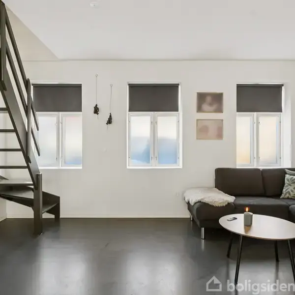 Stue med grå hjørnesofa til højre, et lille rundt bord foran sofaen, og en trappe til venstre. Tre vinduer med sorte rullegardiner slipper lys ind.