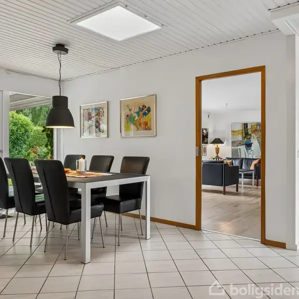 En spisestue med et rektangulært bord og syv sorte stole. Bordet står tæt på store vinduer, der fører ud til en terrasse. De lyse vægge hænger med farverige malerier.
