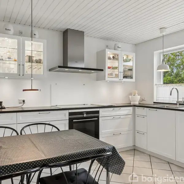 Et hvidt køkken med sorte bordplader, hvor et spisebord med mørkt prikket dug står i forgrunden. Der er et stort vindue, beholdere og et køleskab i baggrunden.