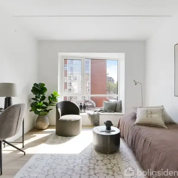 En stue med en brun sofa står op ad væggen. Et rundt sort bord står foran sofaen. Ved siden af står en skrivebordsstol og skrivebord. Store vinduer lader lys strømme ind.