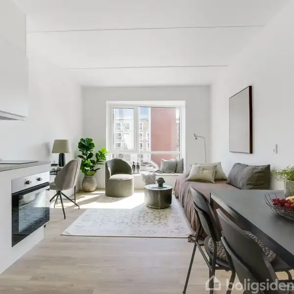 Et moderne køkken-alrum med hvidt interiør. Køkkenet til venstre har en indbygget ovn, og en sort spisebord står til højre med frugt på toppen. En lys stue ses i baggrunden.