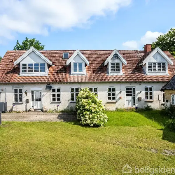 Hvidt hus med rødt tegltag står i sollys. Foran huset er en grøn græsplæne med en busk i midten, omgivet af andre bygninger. Himmel og træer ses i baggrunden.