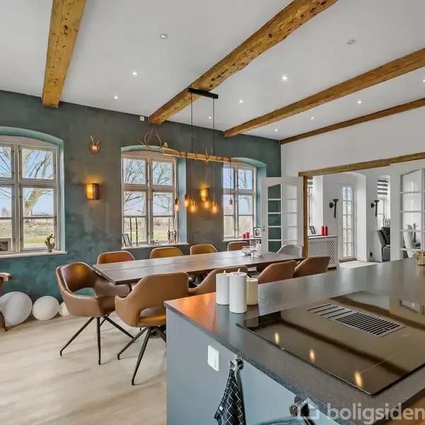 Et stort køkken-alrum har et træbord med stole under en lysarmatur. Loftet er prydet med træbjælker, og store vinduer giver udsigt til træer udenfor.