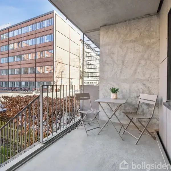 Balkon med to foldestole og et lille rundt bord med en plante. Står i en moderne lejlighedskompleks, omgivet af høje, glas- og murstensbygninger i bymiljø.