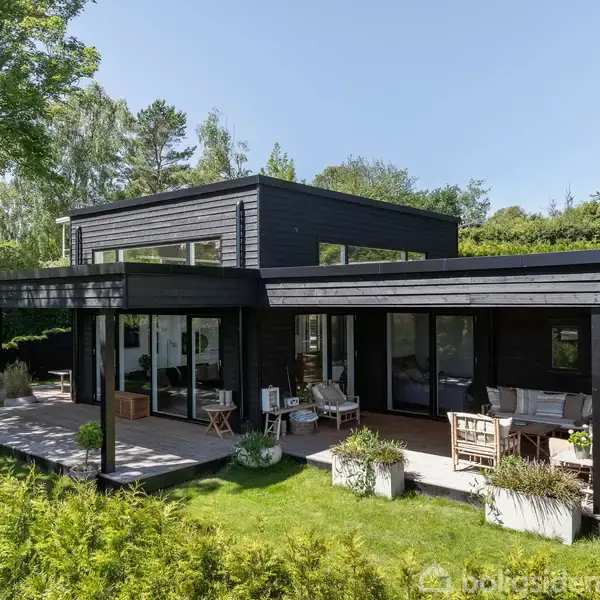Et moderne, sortmalet hus med flade tage står i en frodig have. Store vinduer spejler solen, mens terrassen er møbleret til afslapning. Træer og grønne buske omkranser stedet.