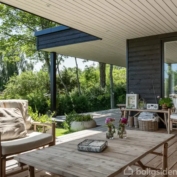 En træterrasse med to komfortable stole og et bord dekoreret med blomster i vaser. Terrassen har et moderne udhæng og er omgivet af frodig grøn natur.