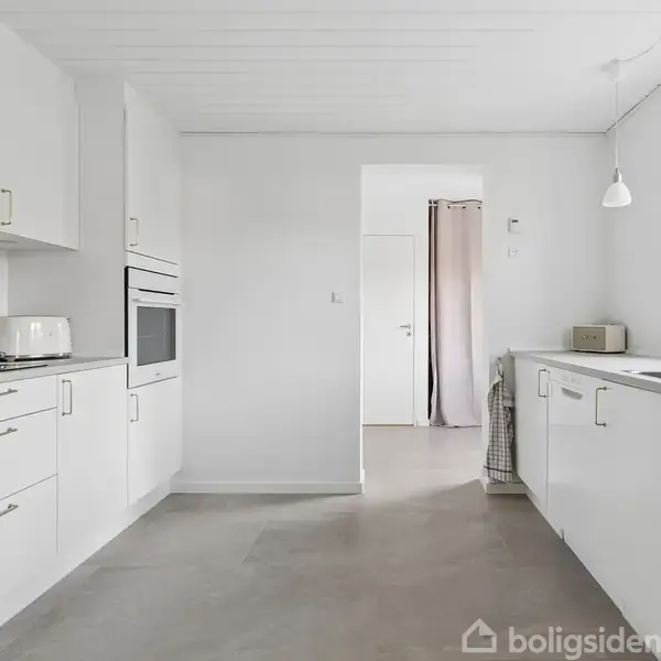Et moderne køkken med hvide skabe og indbyggede apparater; en elkedel står på bordpladen. Rummet er lyst og enkelt med en hvid væg og et vindue mod højre.