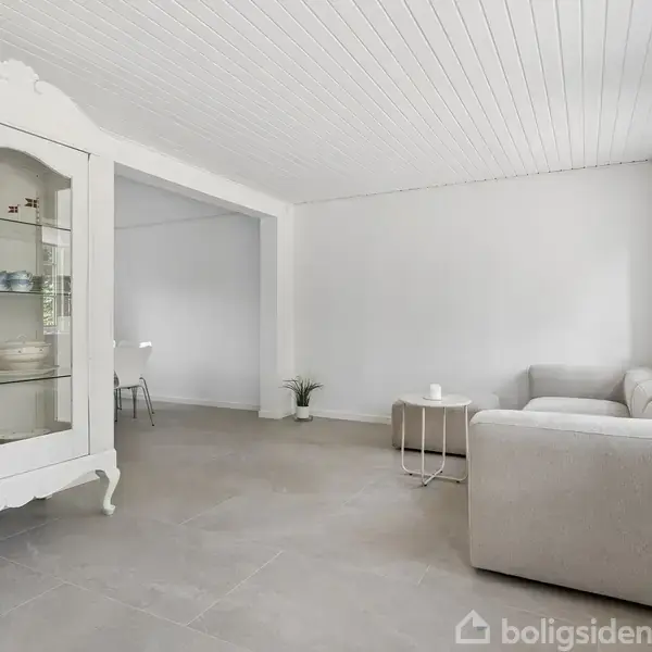 Et hvidt vitrineskab står i en stue. Til højre ses en beige sofa og et lille rundt bord. Rummet er lyst med hvide vægge og store vinduer.