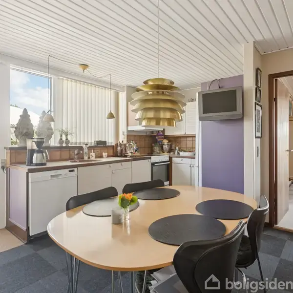 Rundt spisebord med sort dækkeservietter er omkranset af stole. I baggrunden ses et køkken med hvidt interiør og en gylden lampe. Glasdør åbner ud til en terrasse med parasol.