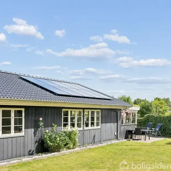 Et sort træhus med solpaneler på taget står i en grøn have. Et flagstang med flag vajer i vinden. Havemøbler ses ved et terrasseområde.