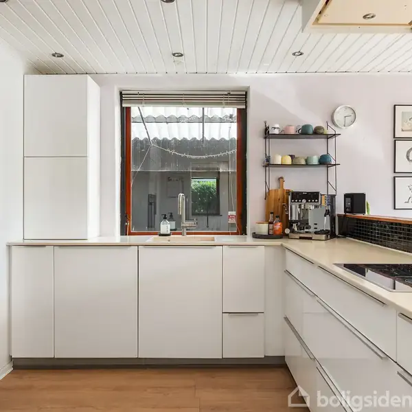 Et moderne køkken med hvide skabe og indbygget ovn. En kogeplade er på højre køkkenbord. Vindue med udsigt til en have. Hylde med kopper og vægudsmykning.