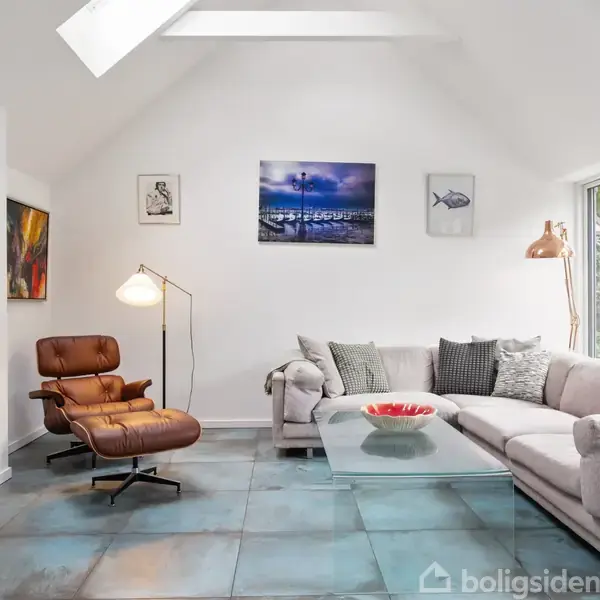 Et rummeligt moderne stueområde med en stor grå hjørnesofa omkring et glasbord. En læderlænestol står ved siden af en stående lampe. Stuen har lyse vægge og kunstværker.