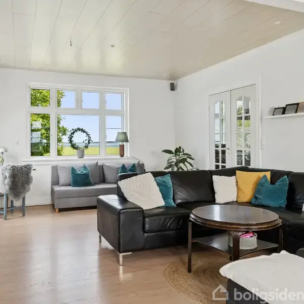 En sort lædersofa arrangeret i en stue med træbord foran. Rummet har lyst trægulv, hvidmalede vægge, store vinduer med udsigt til grøn natur og stilren indretning.