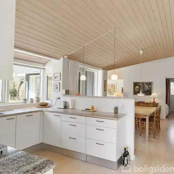 Et moderne køkken med hvide skabe og en grå bordplade, udstyret med husholdningsapparater. Store vinduer slipper naturligt lys ind. Til bagsiden ses en spisestue med træloft og hængelamper.