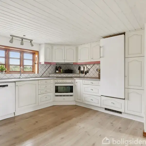 Køkken med hvide skabe og integrerede hvidevarer. Ovn og kogeplader er centreret under et vindue med udsigt. Rummets gulv er lyst træ, og der er et hvidt loft.