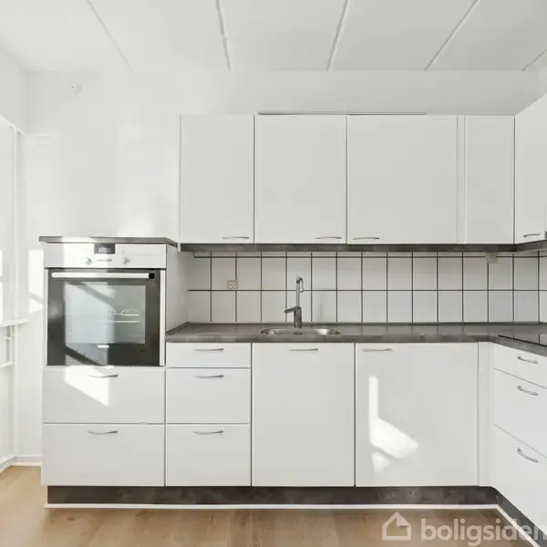 Hvidt køkken med ovn og komfur, integreret i et hjørne. Køkkenet har overskabe og underskabe, en vask, og fliser på væggen. Trægulv ligger i det lyse rum.