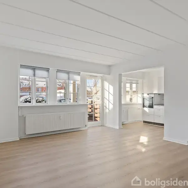 Stue med lyse trægulve og hvide vægge, åbner mod et køkken i baggrunden. Store vinduer og glasdør giver naturligt lys. Radiator under vinduerne.