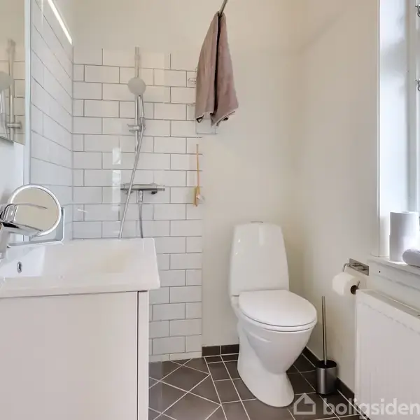 Vask er placeret ved siden af et toilet, med en bruser på væggen bagved. Håndklæde hænger i badeværelset med hvide fliser og et vindue ved siden af. Tootbhurstning ved vasken.