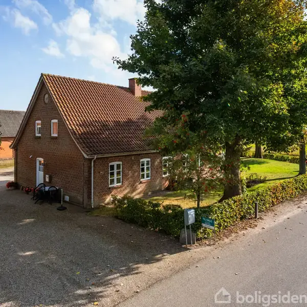 En murstenshus med en rød tag står ved en stille vej. Huset er omgivet af træer og grønne marker under en blå himmel.