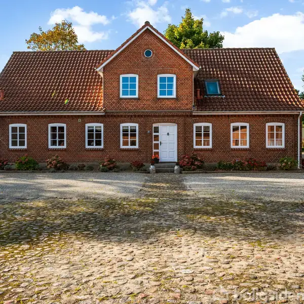 Rødstenshus med symmetrisk facade, står foran en brostensbelagt gårdsplads, omkranset af grønne træer og buske under en blå himmel.
