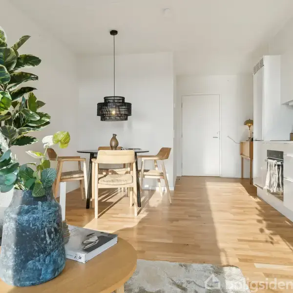 En moderne stue med trægulv har et spisebord og stole i baggrunden. En blå vase med grønne planter står på et rundt sofabord. Køkkenet er til højre.