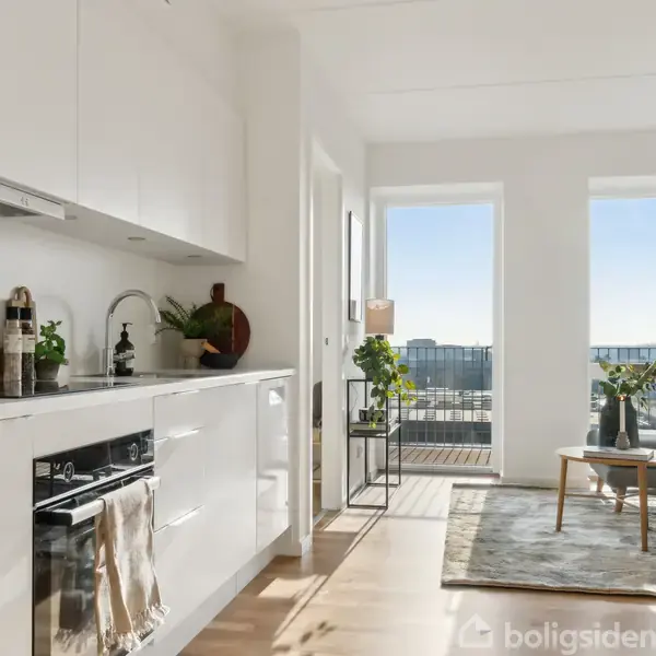 Et lyst køkken med hvide skabe har en sort ovn og planter på bordpladen. I baggrunden ses en stue med sofa, tæppe, sofabord, store vinduer og en balkon.
