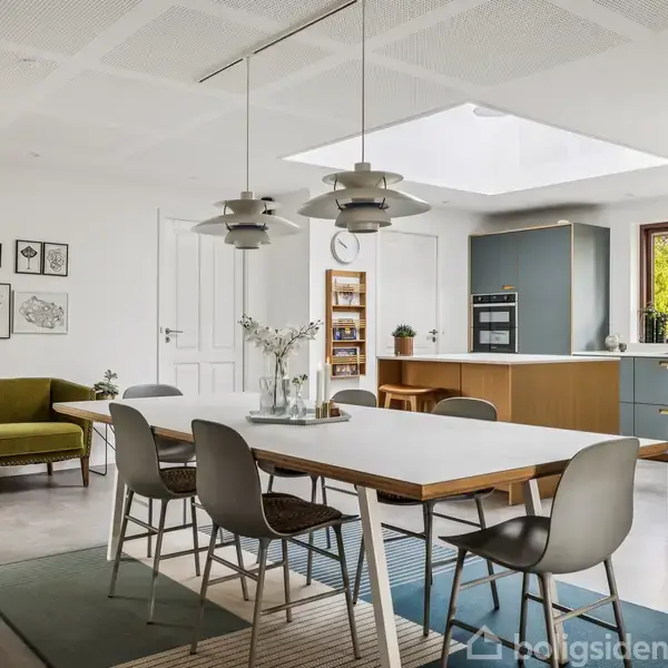 Bord med stole står i et moderne køkkenalrum. Rummet har en grøn sofa, billeder på væggen, og store vinduer. To pendellamper hænger over bordet, og et lysindfald fra ovenlys belyser området.