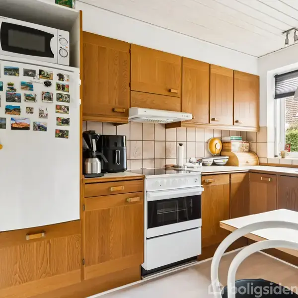 Køkken med træskabslåger, hvid ovn og mikroovn. Køleskabet har farverige magneter. Bordpladen er dekoreret med køkkenudstyr. Vindue med udsigt til grønne buske lyser rummet op.