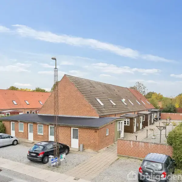 En række røde murstenshuse med skråtag og gavlvinduer står på en stille gade. Biler holder parkeret langs fortovet, og grønne buske omringer området i et roligt beboelseskvarter.