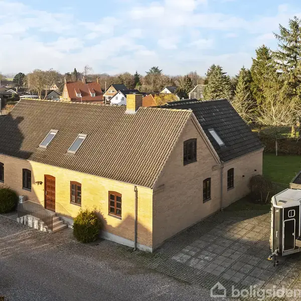 Et stort stenhus med skråt tag og flere vinduer står stille på en grusvej. Byggeriet er omkranset af andre bygninger og grønne træer i et beboelsesområde.