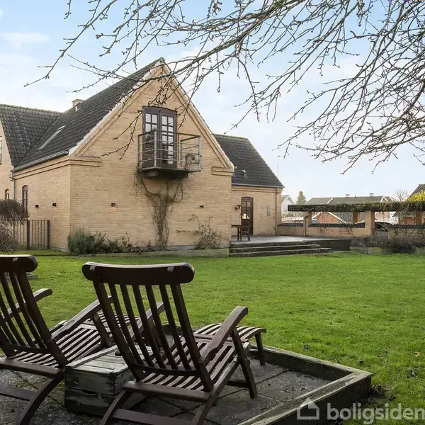 To træstole står i en have på en lille platform. I baggrunden ses et gult murstenshus med en balkon og et grønt græsareal omgivet af træer.
