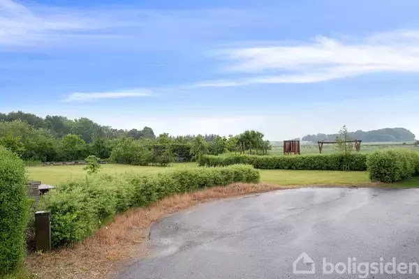 Asfaltvej i forgrunden fører til grønne områder med buske og træer. Blå himmel og skov i baggrunden skaber en fredelig landskabsatmosfære.